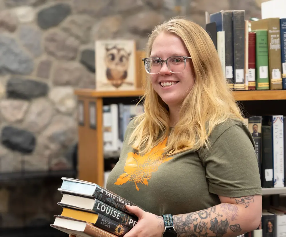 Smiling person in a library holding a stack of books.