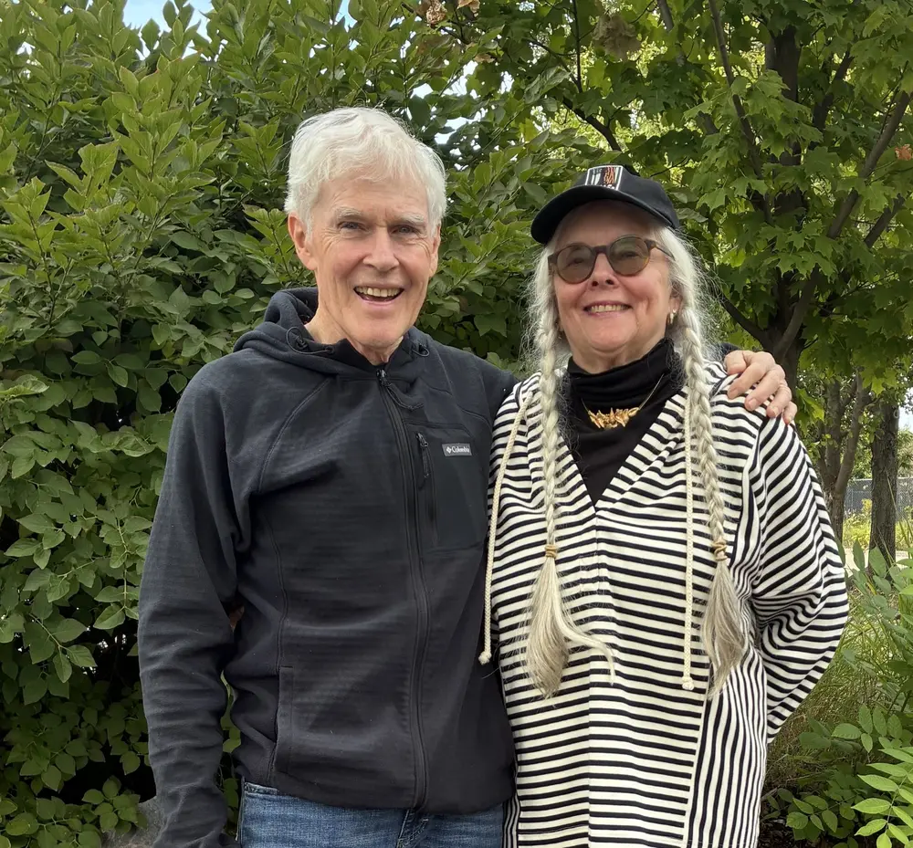 Elderly couple smiling outdoors, surrounded by lush green trees.