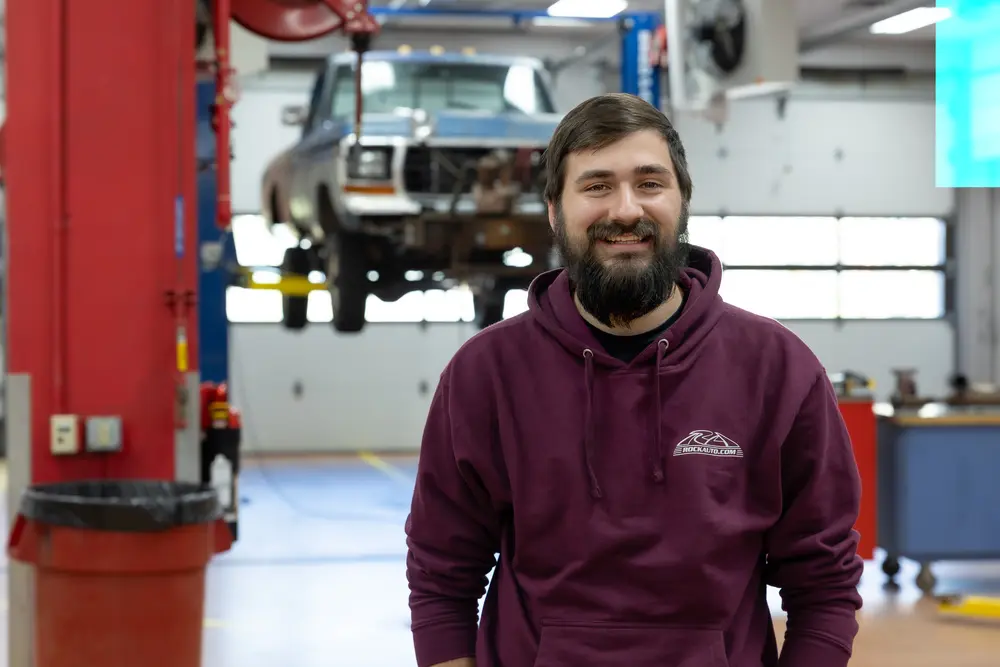 Man in a garage with a vehicle lifted in the background.