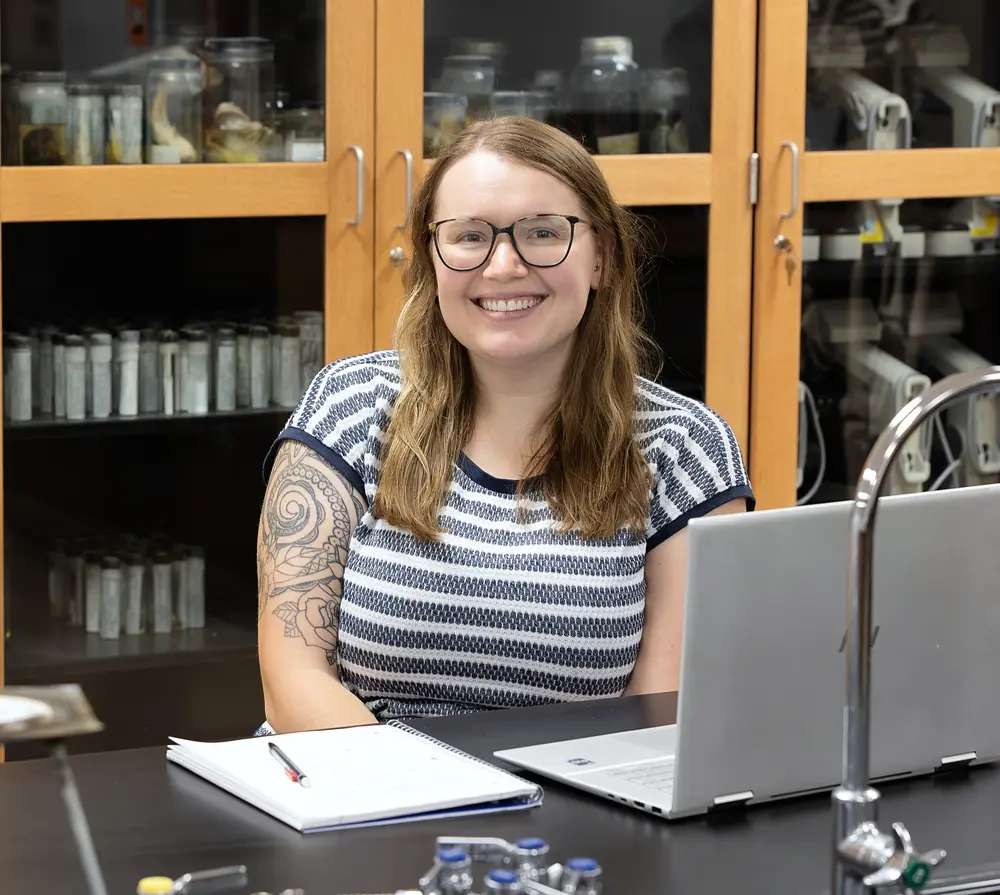 Smiling woman with glasses in a lab, sitting at a desk with a laptop and notebook.