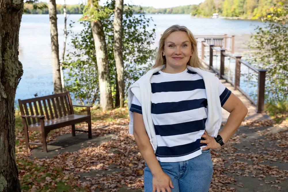 Woman smiling by a lakeside in autumn, wearing striped shirt and jeans.