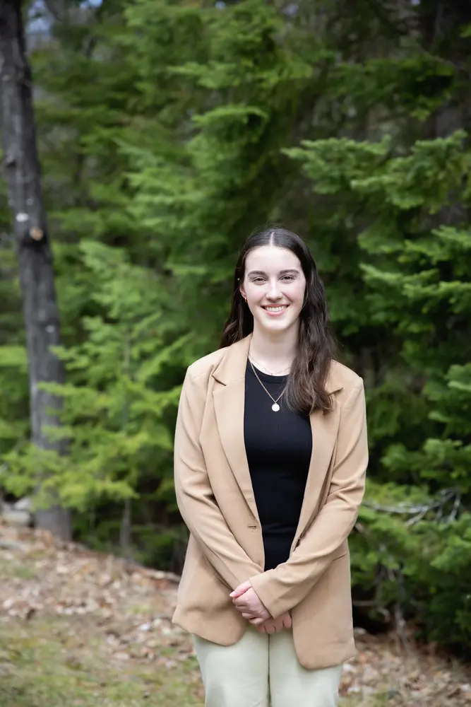 Young woman smiling, standing in a forest, wearing a tan blazer and black top.