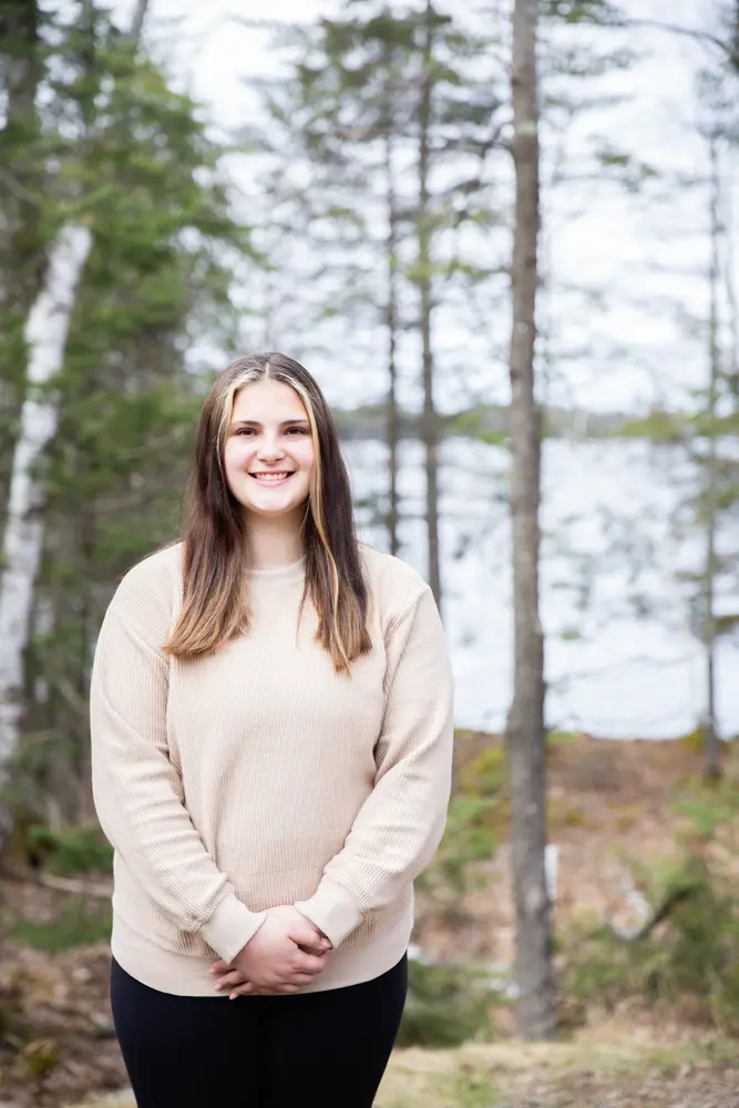 Smiling woman standing in a forest with a lake in the background.