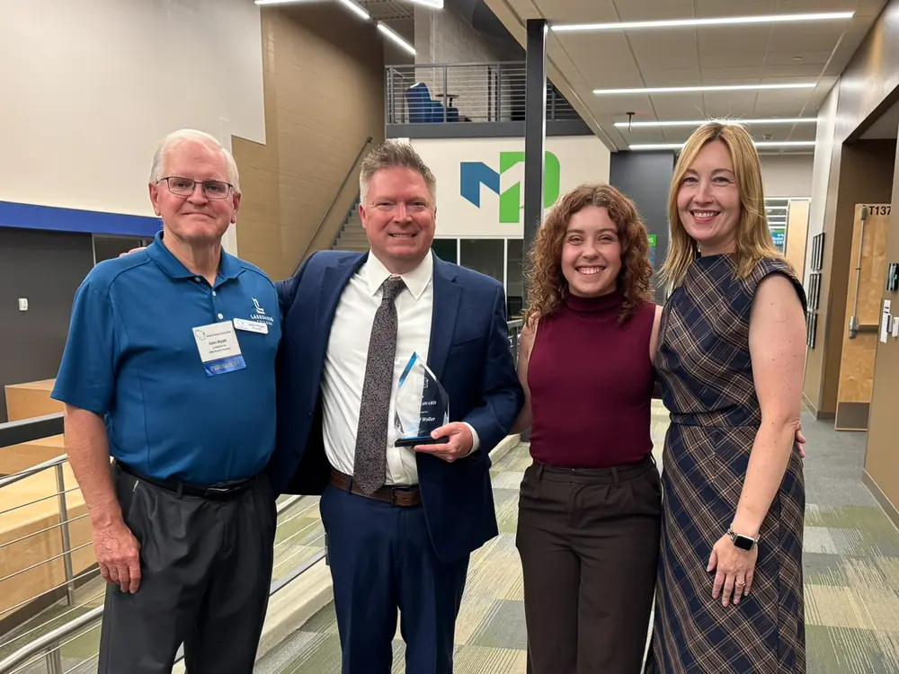 Four people smiling in a well-lit hallway; one holds an award.