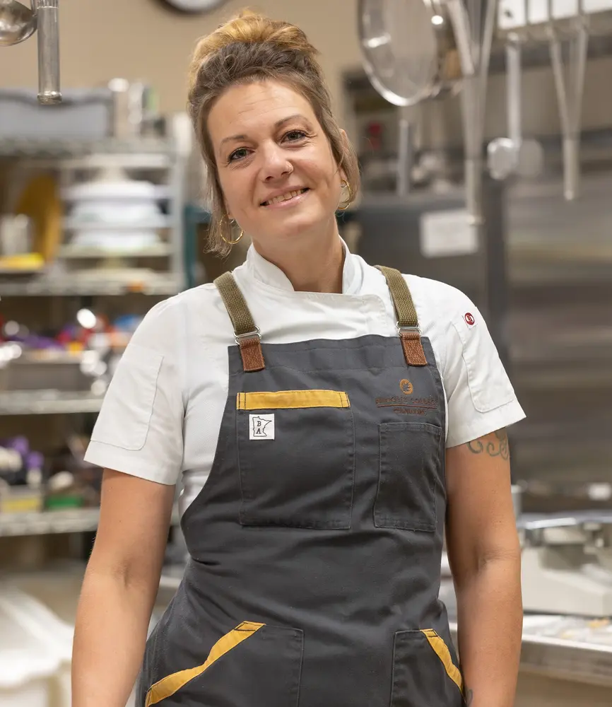 Chef smiling in a kitchen, wearing a black apron over a white shirt.