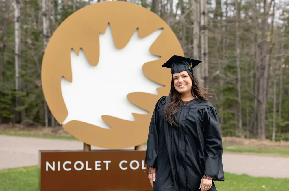 Woman with long brown hair smiling in a graduation cap and gown in front of a large Nicolet College sign.