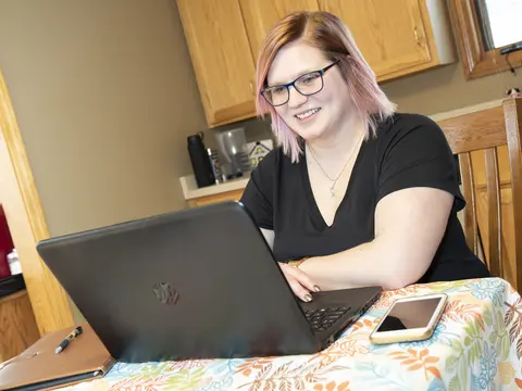 Woman with glasses working on a laptop at a kitchen table, smiling.