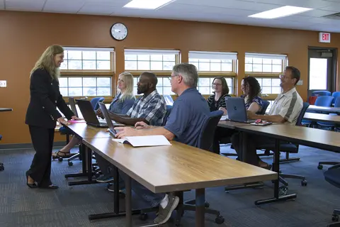 Facilitator at a Grow North meeting talking to six seated adults with laptops in a classroom.