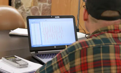 Male student in plaid shirt working on a laptop at a desk with papers and a calculator.