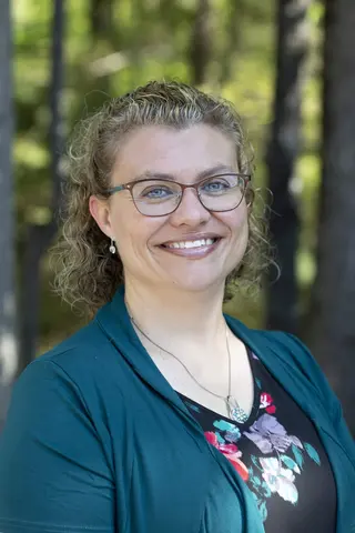 Smiling woman with glasses, curly hair, outdoors in forest setting.