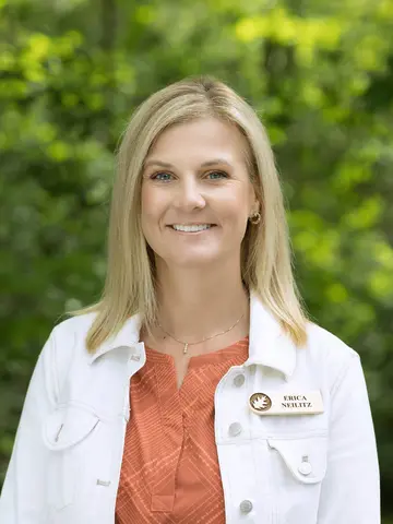 Smiling woman in a white jacket outdoors with greenery.