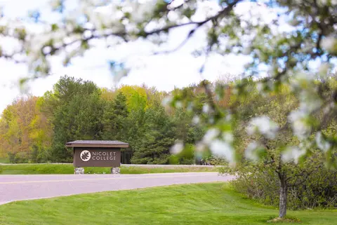 Nicolet College sign in distance, framed by blurred branches and white crab apple blossoms.