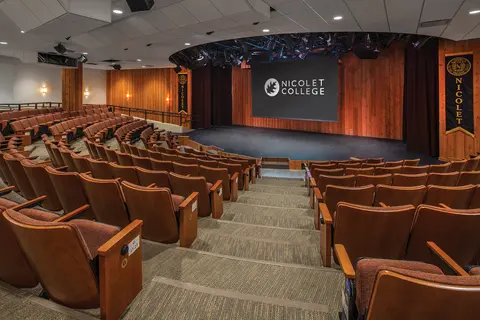 Theatre with rows of brown seats facing a stage and projector screen reading "Nicolet College."