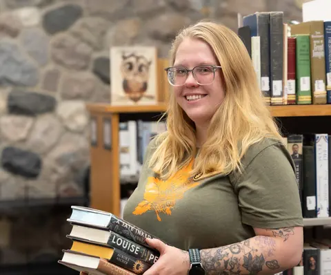 Smiling person in a library holding a stack of books.