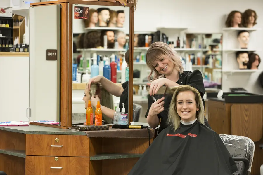 Female cosmetology student styling a woman's hair in a salon, both smiling.