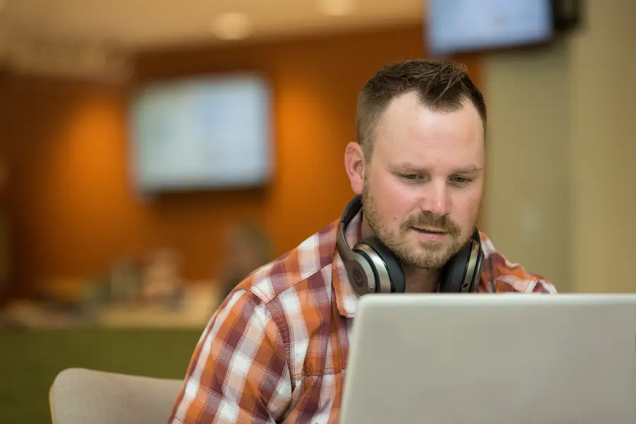 Male student working on laptop, wearing headphones and a plaid shirt.