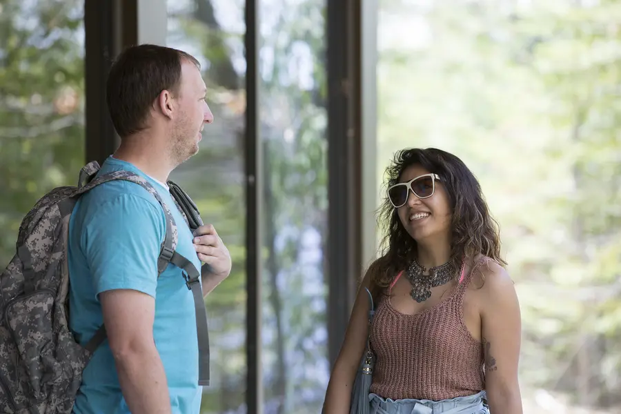 Male student with backpack and female student wearing sunglasses, talking by a window outdoors and smiling.