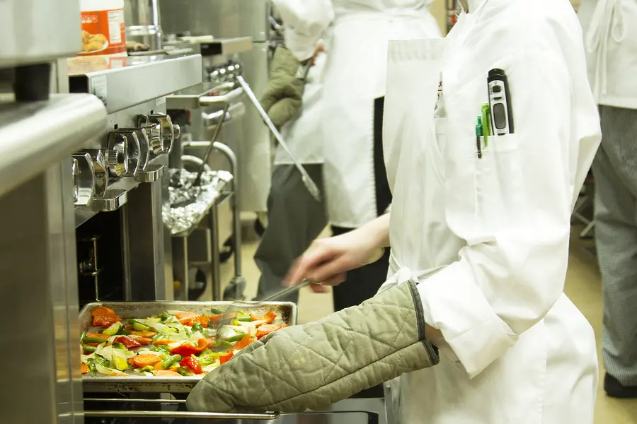 Female culinary student in uniform putting vegetables into an oven in a professional kitchen.