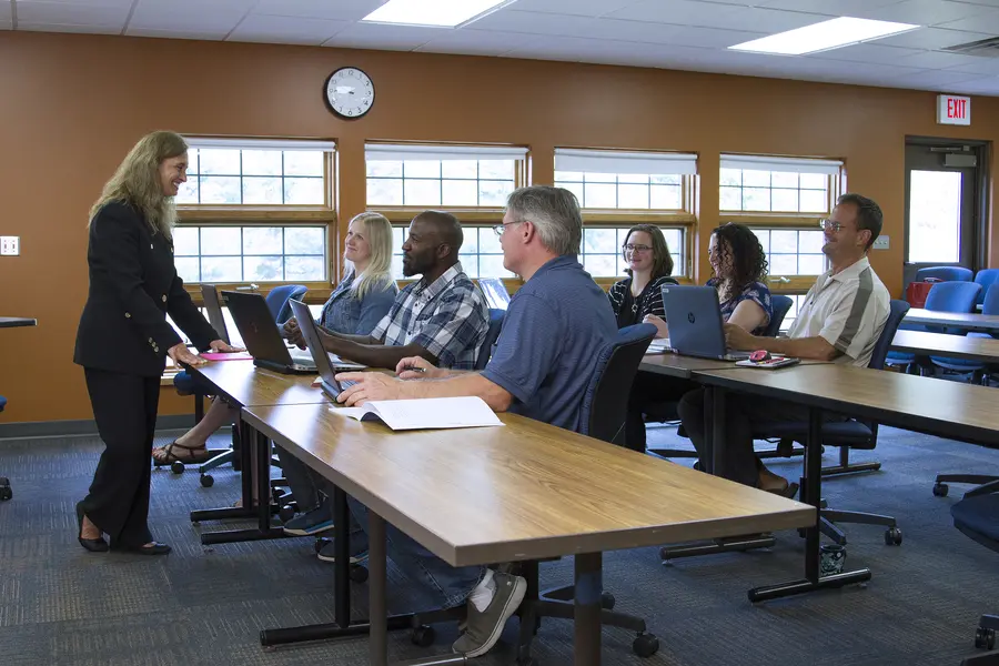 Facilitator at a Grow North meeting talking to six seated adults with laptops in a classroom.