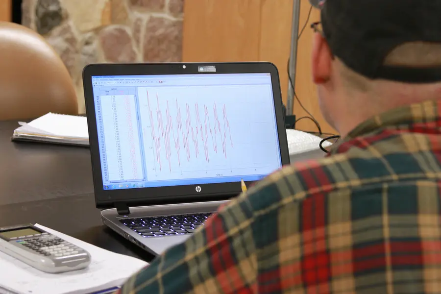 Male student in plaid shirt working on a laptop at a desk with papers and a calculator.