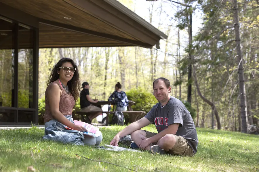 Two students sitting on grass, smiling, trees and a building in background.