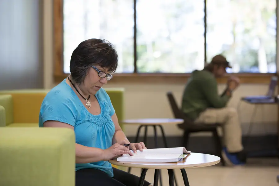 Female student reading at a table in a bright, calm room; another person in background on a laptop.