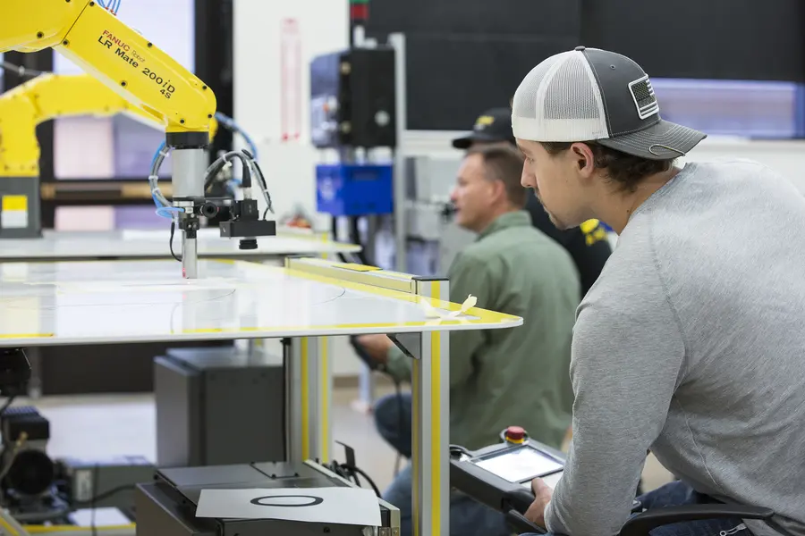 Electromechanical student operating a robotic arm in a workshop setting.