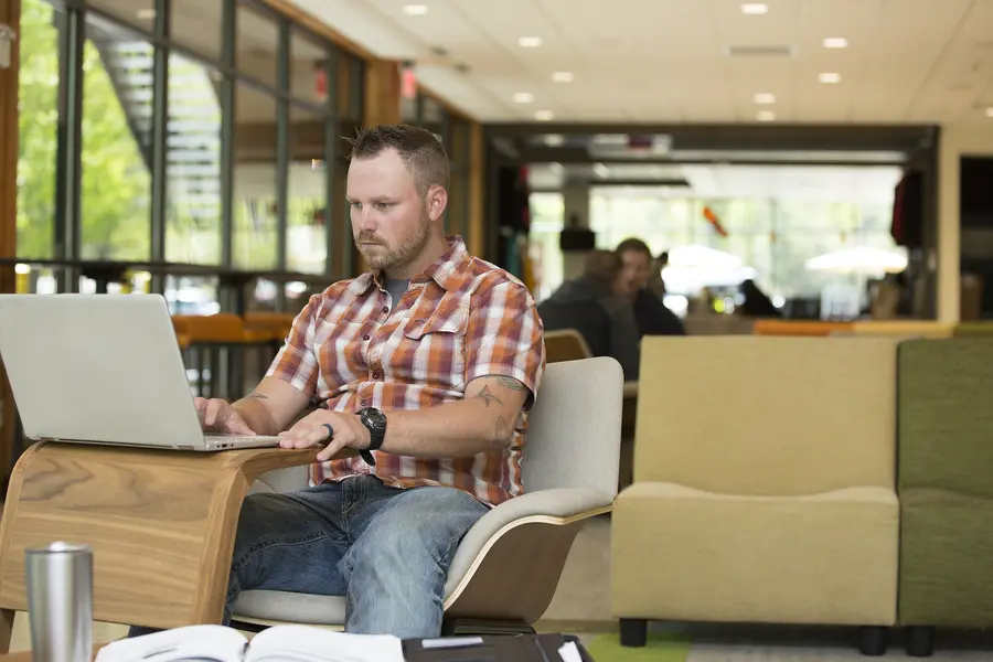 Male student in plaid shirt working on laptop in a modern lounge on campus.