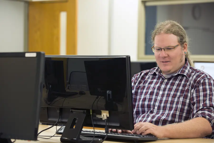 Student with glasses working at dual monitors in an office setting.