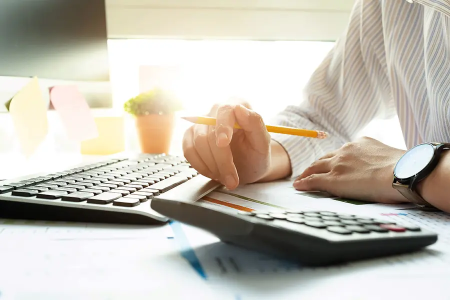 Person using calculator at desk with computer and keyboard.