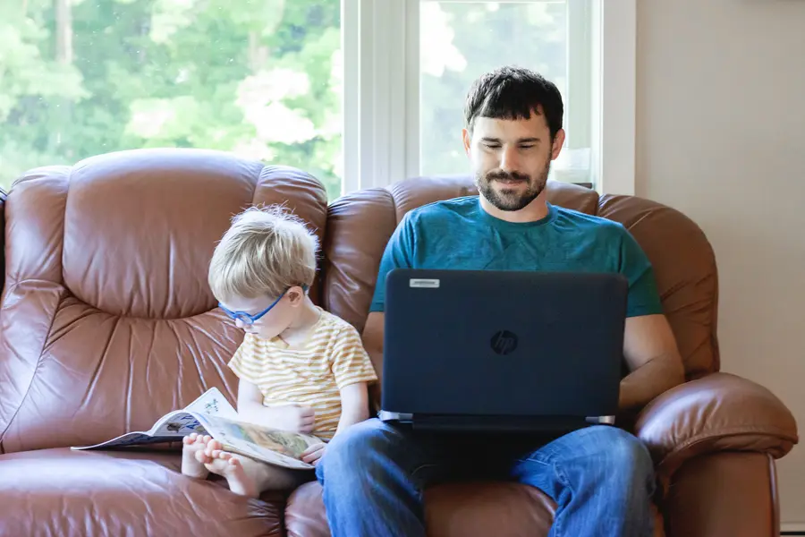 Man using a laptop next to his son reading on a brown sofa, window view of trees.