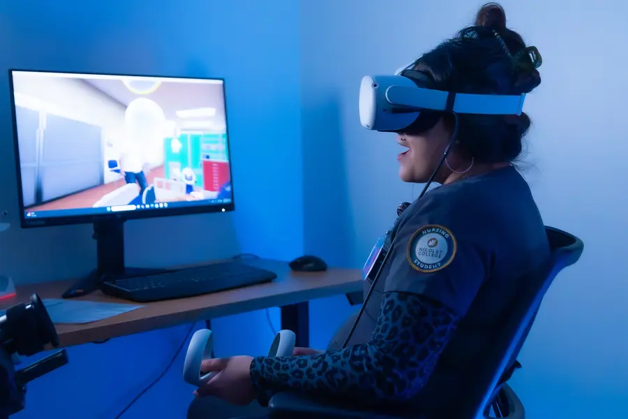 Nursing student using VR headset and controllers, seated by a computer monitor.