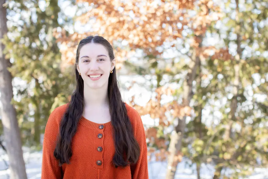 Smiling female scholarship recipient in red sweater outside with autumn trees.