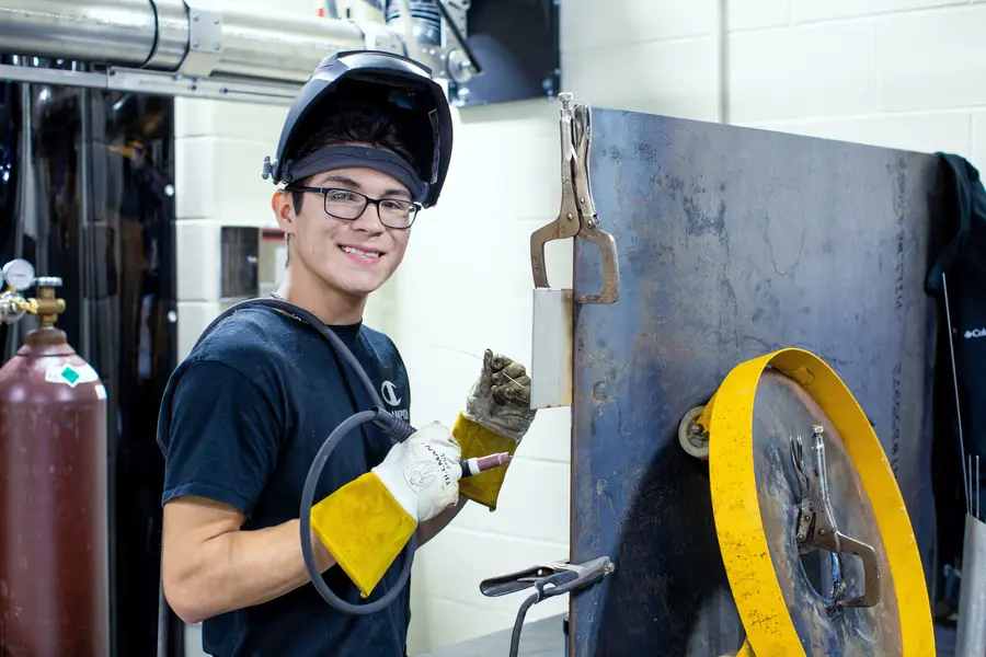 Young welder smiling, holding a welding tool, wearing protective gear.