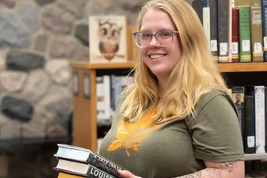 Smiling person in a library holding a stack of books.