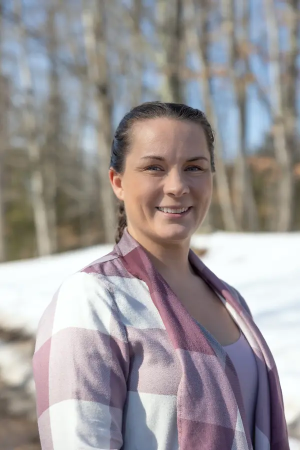 Smiling woman in a plaid shirt stands outside in a snowy forest setting.