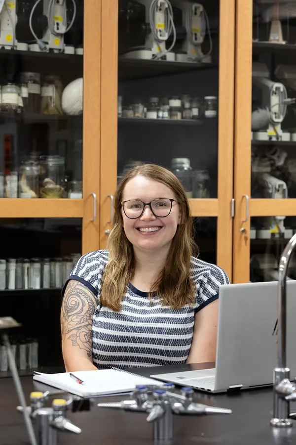 Smiling person with glasses in a science lab, sitting at a laptop.