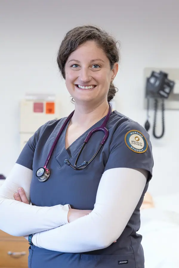 Healthcare worker smiling in a medical room, arms crossed, wearing scrubs and a stethoscope.