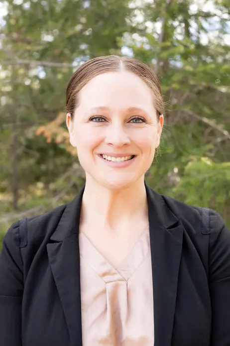 Smiling woman in blazer, outdoors with trees in background.
