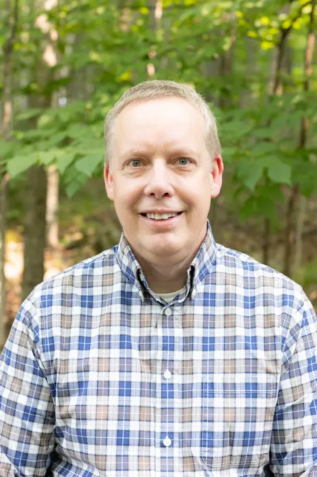 Smiling man in checkered shirt, standing in a leafy forest.