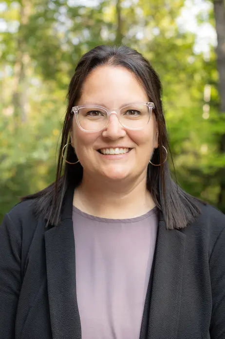 Smiling person in glasses and blazer, standing outdoors with trees in the background.