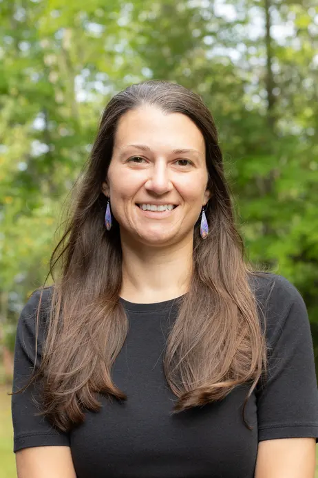 Smiling woman with long hair, wearing a black shirt, standing outdoors with trees behind.