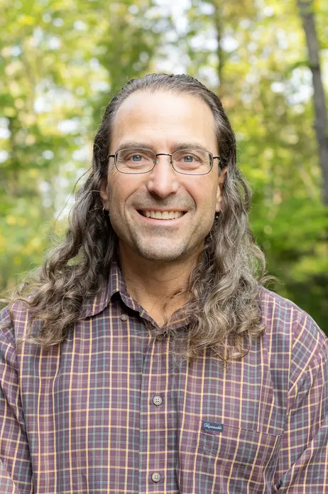 Man with long hair and glasses smiling in a forest setting.