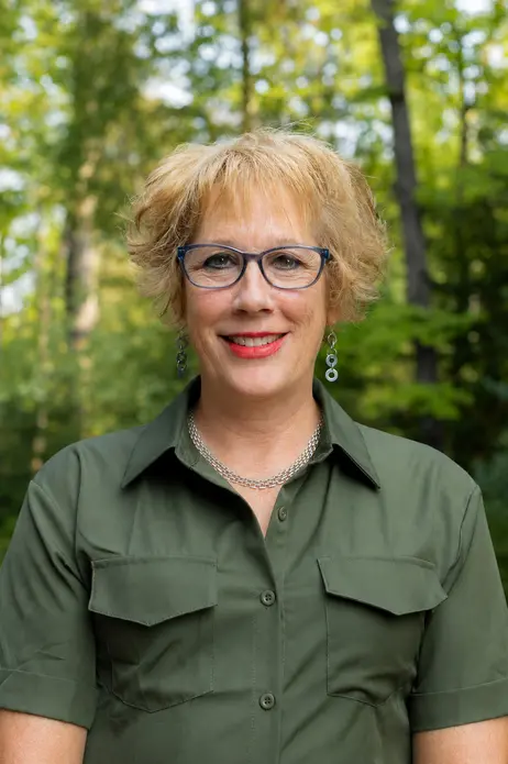 A person with short blond hair, wearing glasses and a green shirt, smiles outdoors.