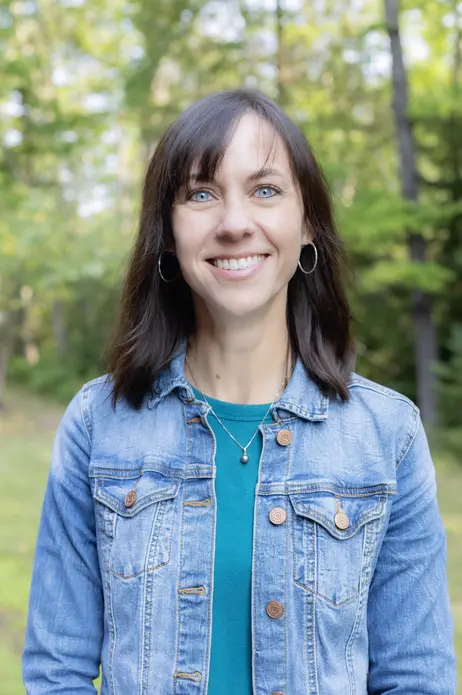 Smiling woman in a denim jacket outdoors with green trees.