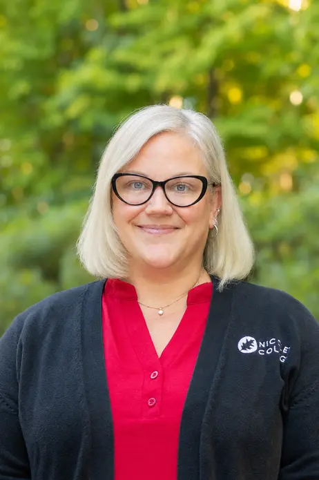 A person with glasses and light hair, wearing red and black, smiles outdoors.